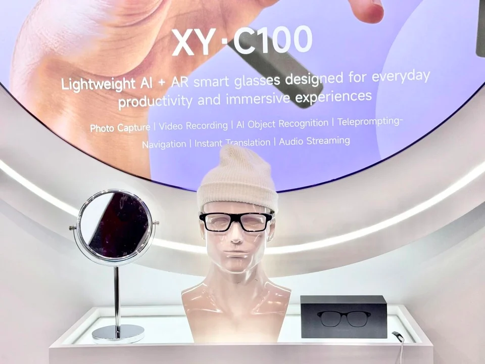 Close view of a mannequin wearing C100 glasses and a white hat, beside a mirror and product box under a purple XY-C100 display